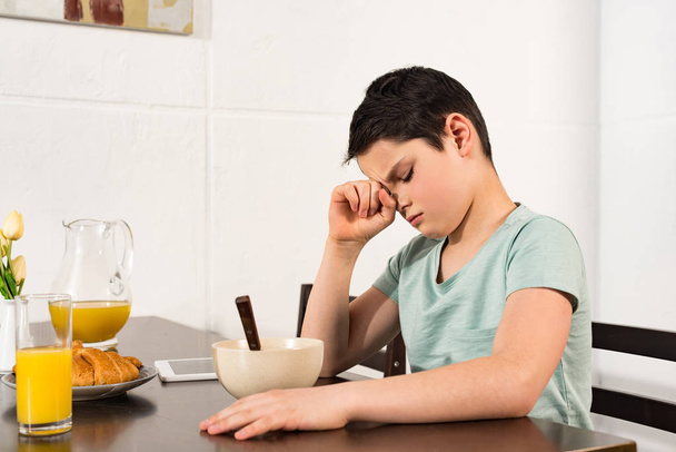 triest jongen zittend aan tafel tijdens het ontbijt in de keuken - Foto, afbeelding
