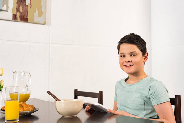 χαμογελαστή αγόρι χρησιμοποιώντας ψηφιακό δισκίο κατά τη διάρκεια του πρωινού στην κουζίνα - Φωτογραφία, εικόνα