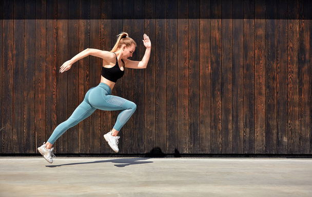 Young European woman sports model posing outdoors along wooden wall, running, jumping, jogging, stretching, wearing in sport closes, sneakers and black top - Photo, Image