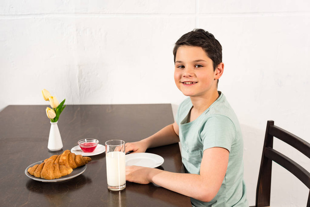 usměvavý chlapec sedící u stolu s croissanty, sirupem a skleněnými mlékem - Fotografie, Obrázek