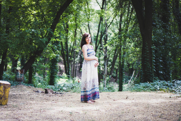 Piękny portret kobiety w ciąży w lekkiej sukni w kolorowym letnim lesie. Pojęcie ciąży i pory roku. Obróbka filmowa. - Zdjęcie, obraz