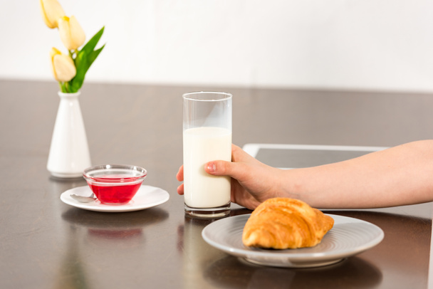 cropped view of boy sitting at table with croissant, syrup and glass of milk - Фото, изображение