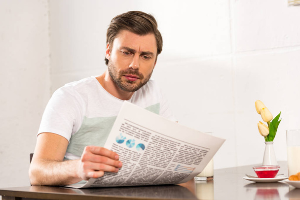 focused bearded man reading newspaper during breakfast in kitchen - Photo, Image