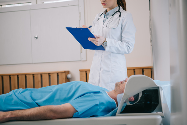 vue recadrée du jeune radiologue écrivant sur le presse-papiers alors qu'il se tenait près du patient allongé sur le lit du scanner IRM
 - Photo, image