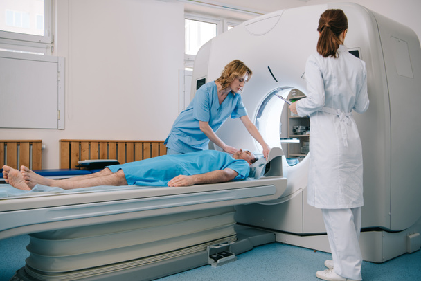 radiologista v bílém plášti stojící poblíž CT Scanner, zatímco ošetřovatelka připravuje pacienta na diagnostiku - Fotografie, Obrázek