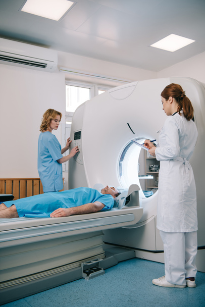 radiolog piśmie w schowku, podczas gdy asystent pracy skaner CT w pobliżu pacjenta leżącego na skanera łóżko - Zdjęcie, obraz