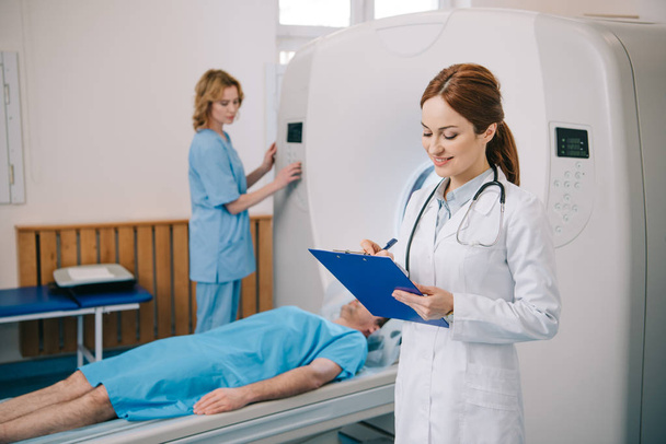 Selektywny fokus uśmiechnięty radiolog piśmie w schowku, podczas gdy asystent pracy skaner CT w pobliżu pacjenta leżącego na CT skanera łóżko - Zdjęcie, obraz