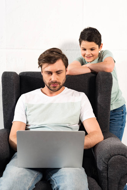 усміхнений син дивиться, як батько використовує ноутбук у вітальні
 - Фото, зображення