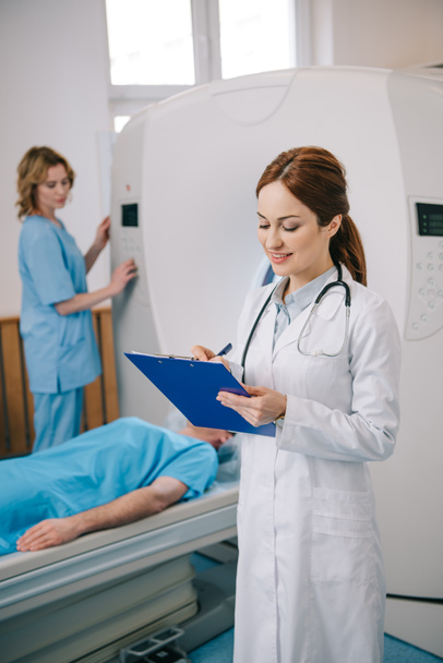 Selektywny fokus radiolog piśmie w schowku, podczas gdy asystent obsługi MRI maszyny w pobliżu pacjenta - Zdjęcie, obraz