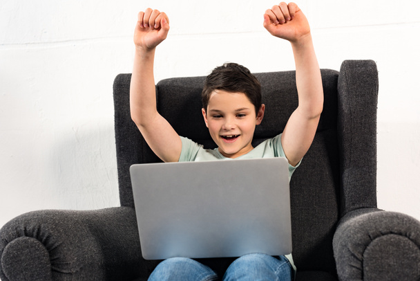 opgewonden jongen in jeans zittend in fauteuil en met behulp van laptop - Foto, afbeelding