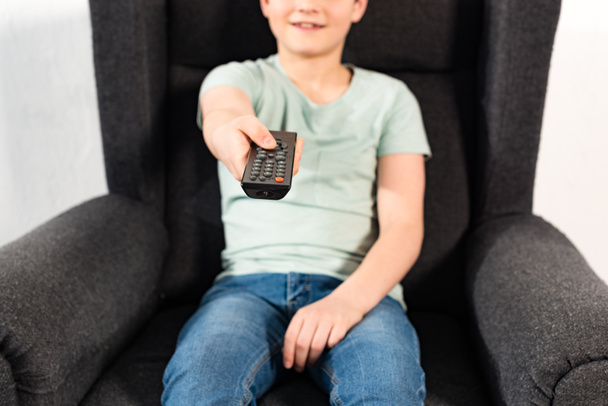 niño en jeans sentado en sillón y sosteniendo mando a distancia
 - Foto, Imagen