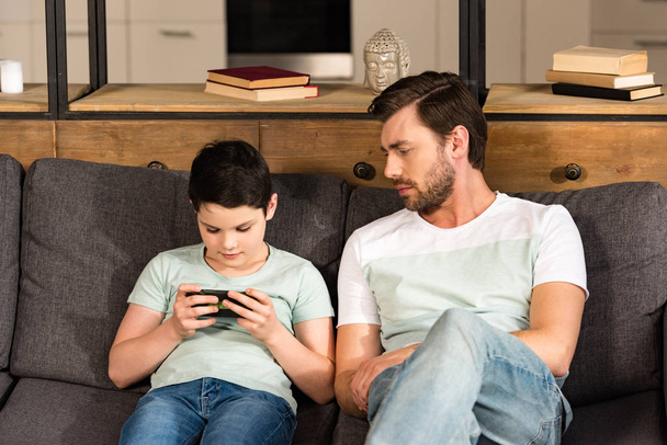 dad looking at son using smartphone while sitting on sofa in living room - Fotó, kép