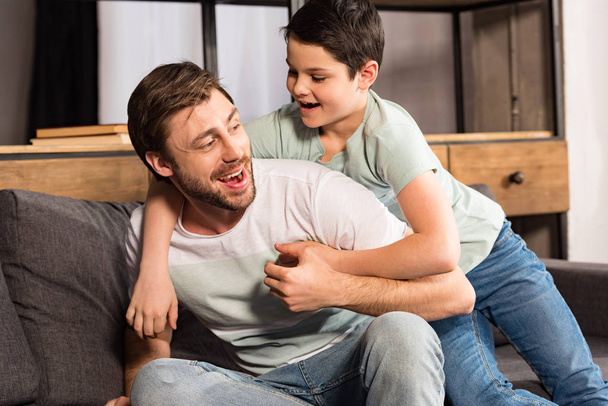 laughing son and dad sitting on sofa and having fun in living room - Fotó, kép