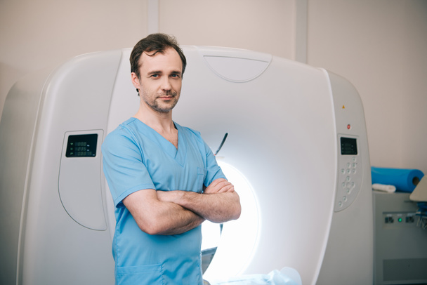 Ένας χαμογελαστός ακτινολόγος στέκεται κοντά σε αναλυτή αξονικής τομογραφίας με σταυρωτά χέρια και κοιτάζοντας την κάμερα - Φωτογραφία, εικόνα