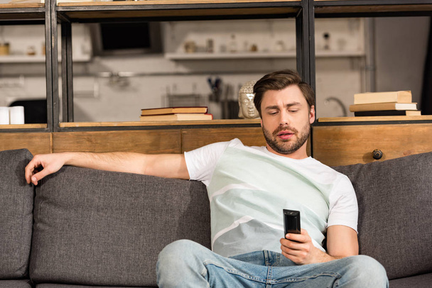 bearded man in jeans sitting on sofa and holding remote controller - Photo, Image