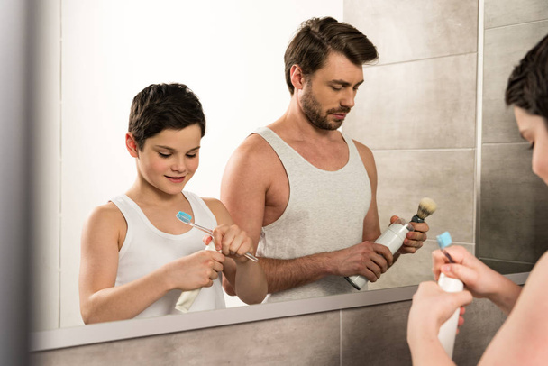 son brushing teeth and dad shaving in morning in bathroom - Photo, Image