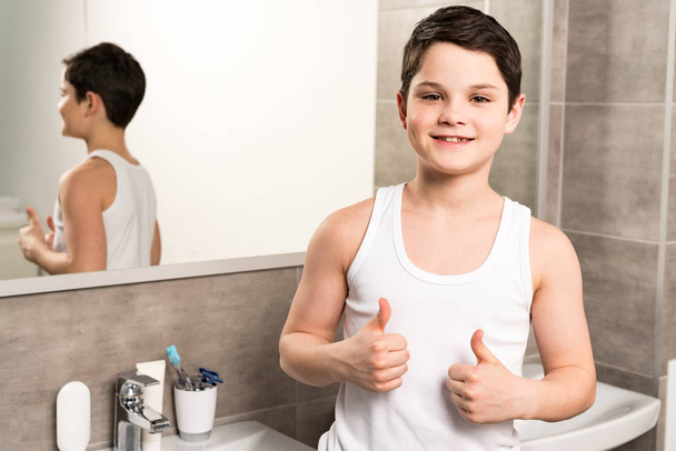 uśmiechnięty chłopak stojący w pobliżu lustro w łazience i pokazując kciuki w górę - Zdjęcie, obraz