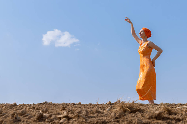 Ethnic Woman in Turban Dances on Clean Soil on Sky and Clouds Background - Foto, imagen