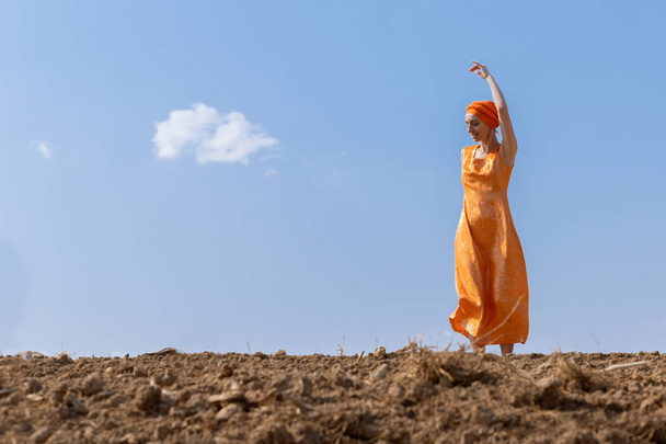 Ethnic Woman in Turban Dances on Clean Soil on Sky and Clouds Background - Фото, изображение