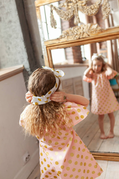 Little pretty girl in a pink dress looks in the mirror at her reflection - Foto, afbeelding
