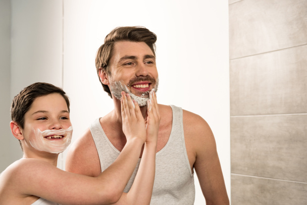 preteen boy applying shaving foam on smiling father in bathroom - Photo, image