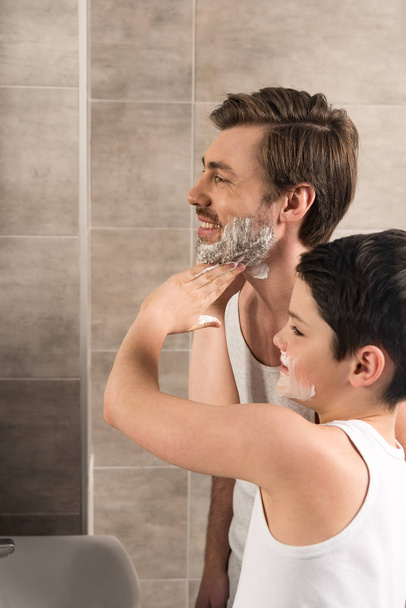 niño preadolescente aplicando espuma de afeitar en el padre sonriente en el baño
 - Foto, Imagen