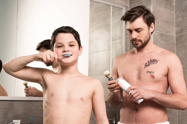 preadolescente niño cepillarse los dientes y padre celebración de espuma de afeitar y cepillo en el baño
 - Foto, Imagen