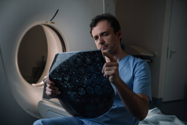 внимательный врач, изучающий диагноз томографии, сидя рядом со сканером КТ
 - Фото, изображение