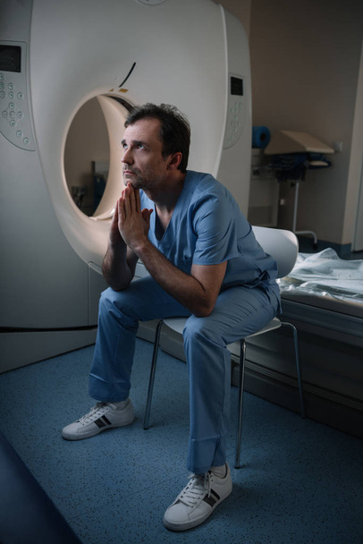 病院でコンピュータ断層撮影スキャナの近くに座って、見上げる深刻な医師 - 写真・画像