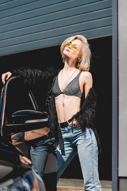 beautiful fashionable young woman in glasses posing near car and garage - Фото, изображение
