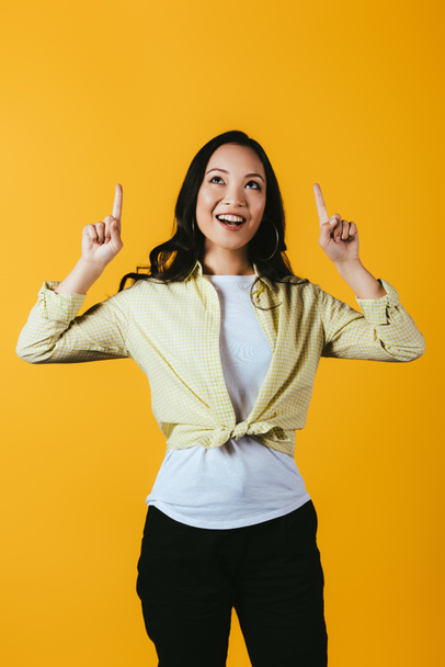 beautiful smiling asian girl pointing up isolated on yellow - Фото, изображение