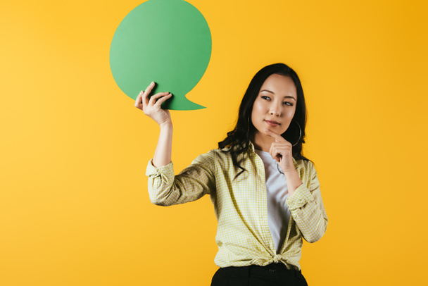 thoughtful asian woman holding green speech bubble, isolated on yellow - Fotó, kép