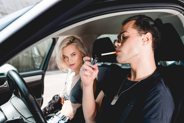 車の中で美しい女の子と座っている間、スタイリッシュなハンサムな男はタバコを点灯 - 写真・画像