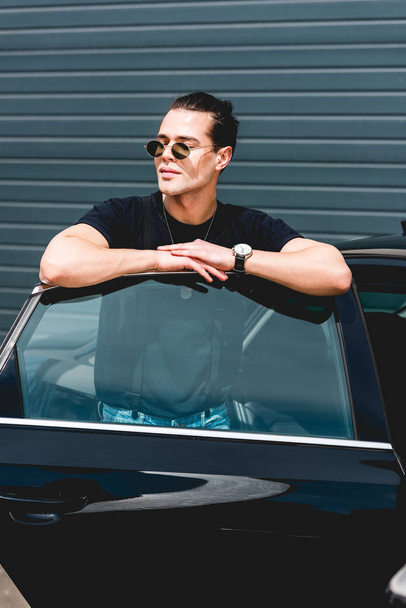 handsome stylish man in sunglasses posing near car and looking away - Zdjęcie, obraz