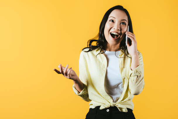 excited asian girl talking on smartphone isolated on yellow  - 写真・画像