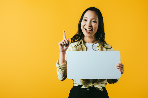 smiling asian girl holding speech bubble and pointing up, isolated on yellow  - Foto, imagen