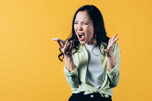 aggressive asian girl screaming on smartphone isolated on yellow  - Foto, imagen
