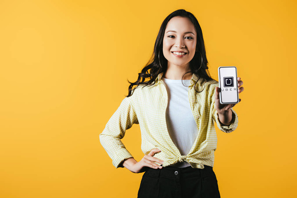 KYIV, UKRAINE - 16 de abril de 2019: menina asiática sorridente mostrando smartphone com aplicativo uber, isolado em amarelo
 - Foto, Imagem