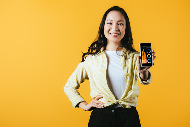 ευτυχισμένο κορίτσι της Ασίας δείχνει smartphone με ενημερωτικό γραφικό, απομονώνεται σε κίτρινο - Φωτογραφία, εικόνα