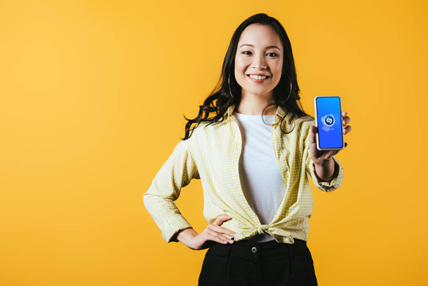 KYIV, UKRAINE - APRIL 16, 2019: smiling asian girl showing smartphone with shazam app, isolated on yellow - Photo, Image