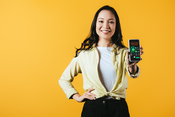 glückliche Asiatin zeigt Smartphone mit Marketinganalyse-App, isoliert auf gelb - Foto, Bild