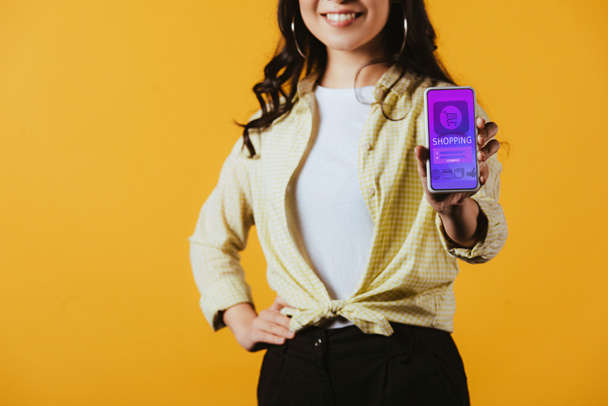 bijgesneden weergave van brunette meisje toont smartphone met shopping app, geïsoleerd op geel - Foto, afbeelding