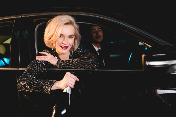 stijlvolle man en mooie glimlachende jonge vrouw in formele slijtage met Champagne fles zitten in de auto - Foto, afbeelding