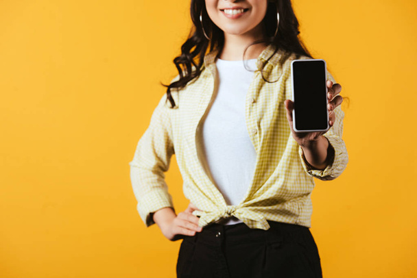 cropped view of smiling girl showing smartphone with blank screen, isolated on yellow - Fotó, kép