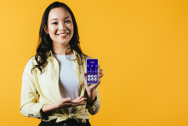 happy asian girl presenting smartphone with health app, isolated on yellow - Photo, Image