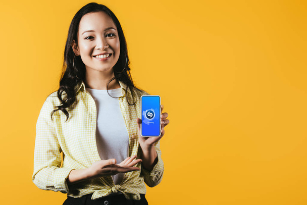KYIV, UKRAINE - 16 de abril de 2019: menina asiática sorridente apresentando smartphone com aplicativo shazam, isolado em amarelo
 - Foto, Imagem
