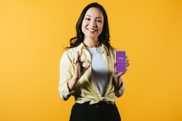KYIV, UCRANIA - 16 DE ABRIL DE 2019: chica asiática sonriente mostrando signo de ok y teléfono inteligente con aplicación instagram, aislado en amarillo
 - Foto, Imagen