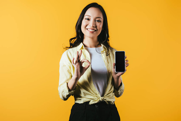 attractive asian girl showing ok sign and smartphone with blank screen isolated on yellow - Photo, Image