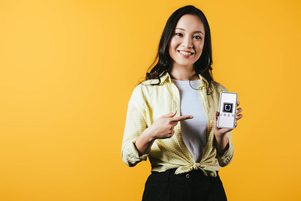 KYIV, UKRAINE - APRIL 16, 2019: attractive smiling asian girl pointing at smartphone with uber app, isolated on yellow - Photo, Image