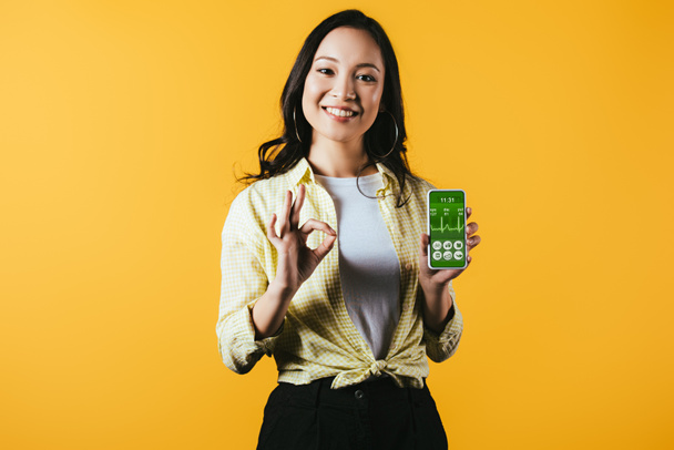 smiling asian girl showing ok sign and smartphone with health app, isolated on yellow - Photo, Image